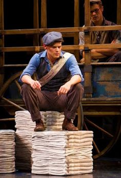 a man sitting on top of stacks of paper next to another man in front of a wagon