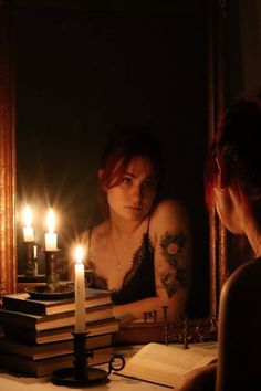 a woman sitting in front of a mirror next to a stack of books with candles