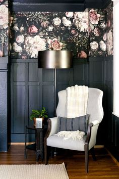 a white chair sitting next to a lamp on top of a hard wood floor covered in floral wallpaper