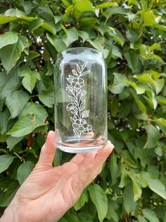 a hand holding up a glass jar with flowers etched on it in front of green leaves