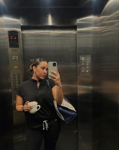 a woman taking a selfie with her cell phone in an elevator holding a coffee cup