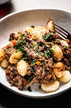 a white bowl filled with meat and potatoes on top of a table next to a fork