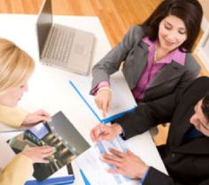 three people sitting at a table with laptops and papers