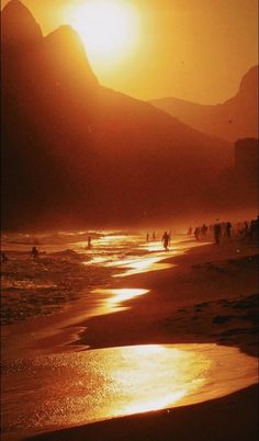 people are walking on the beach as the sun goes down in the mountains behind them