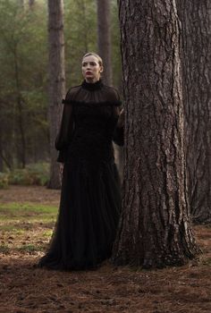 a woman standing next to a tree in a forest wearing a black dress with sheer sleeves