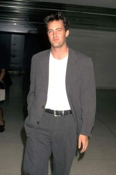 a man in a suit and white t - shirt is standing with his hands in his pockets