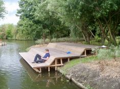 a person sitting on a wooden dock in the water near some trees and grass,