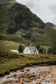 a white house sitting on the side of a lush green hillside next to a river