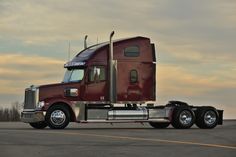 a red semi truck parked on top of a parking lot next to a field and trees