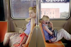 two people sitting on a train with their heads turned to the side, and one person holding a cell phone