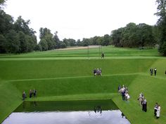 many people are walking around in the grass near a body of water and some trees