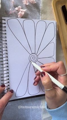 a woman is drawing on a notebook with white paper and black marker, while another person holds a pen in their hand