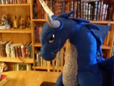 a stuffed blue dragon sitting on top of a wooden table in front of bookshelves