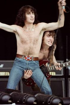 two shirtless men playing guitars on stage with one holding his arms in the air