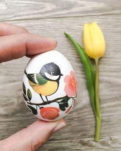 a hand holding an egg with a bird painted on it next to a yellow tulip