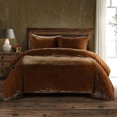 a bed with brown comforter and pillows in a room next to a lamp on a dresser