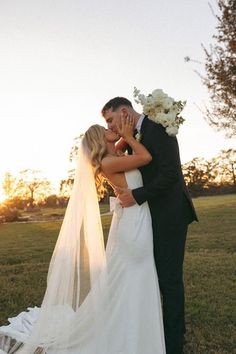 a bride and groom kissing in front of the setting sun at their wedding ceremony, with veil blowing in the wind