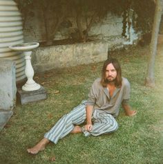 a man sitting on the grass in front of a building with his legs crossed and long hair tied back