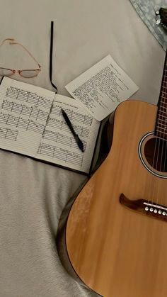 an acoustic guitar and music sheets are on a bed with eyeglasses next to it