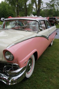 an old pink and white car is parked on the grass in front of other cars