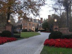 a driveway with red flowers in front of a large brick building and gated entrance