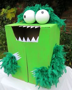 a green cardboard box with two eyes and some feathers on the outside, in front of bushes