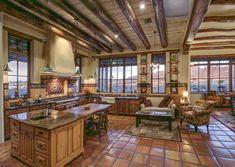 a large open kitchen and living room area with tile flooring, exposed wood beams, windows, and an island in the center