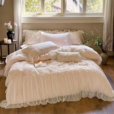 a bed with white ruffles and pillows in front of a window next to a night stand