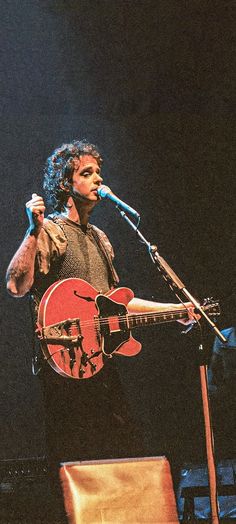 a man holding a red guitar while standing next to a microphone on top of a stage