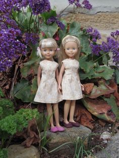 two dolls standing next to each other in front of purple flowers and plants on the ground