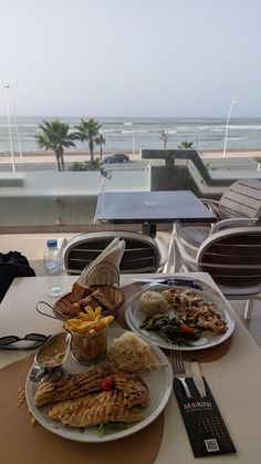 a table with plates of food on top of it near the ocean and beach in the background