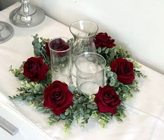 red roses and greenery are arranged in a circle on a white tablecloth with candles