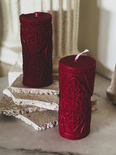 two red candles sitting next to each other on top of a stone slab in front of a window