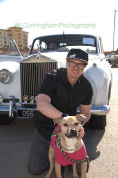 a man kneeling down next to a dog wearing a pink shirt and black baseball cap