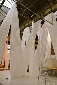 the interior of an art gallery with white drapes hanging from the ceiling and ladders on the floor