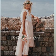 a beautiful young blonde woman standing next to a brick wall wearing a white dress and flower crown