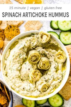 a white bowl filled with spinach artichoke hummus surrounded by crackers and cucumbers