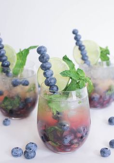 three glasses filled with blueberries, lime and mint sprits on a white surface