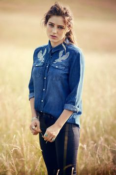 a woman standing in the middle of a field with her hands in her pockets and looking at the camera