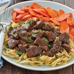 a white plate topped with meat and pasta next to carrots on a wooden table