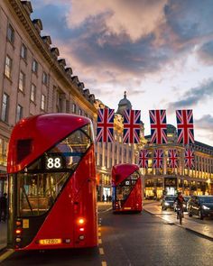 two red double decker buses driving down a street next to tall buildings with flags on them