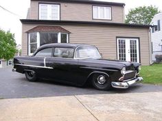 an old black car is parked in front of a house with a brown siding and two windows