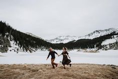 two women are running in the snow holding hands and looking at each other with mountains in the background