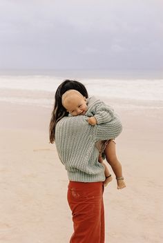 a woman holding a baby doll on the beach in front of an ocean and cloudy sky