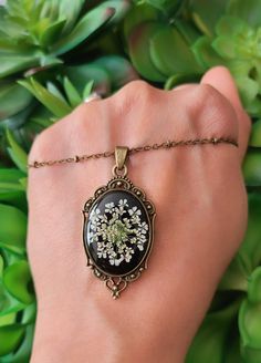 a woman's hand holding a necklace with white flowers on it and green leaves in the background