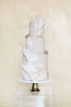 a three tiered cake with white frosting and flowers on the top is sitting on a glass stand