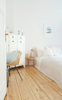 a bedroom with white walls and wooden flooring is pictured in this image, there is a wicker chair next to the bed