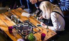 a group of people sitting around a wooden table with cards and drinks on top of it