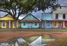 colorful houses are reflected in a puddle on the dirt ground near a tree and grass field