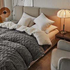 a bed with pillows and blankets on top of it next to a night stand, chair and lamp